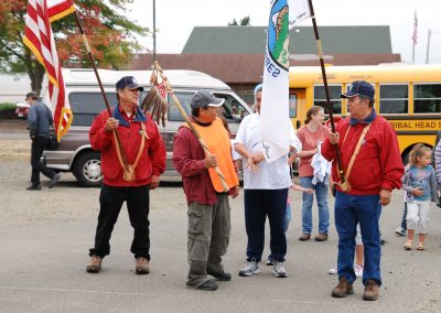Tribal members standing in Siletz before Run to the Rogue. From left to right, they are holding: the American flag, the Eagle Staff, and the CTSI flag.