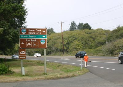 A Tribal member walking down the side of the road holding the Eagle Staff. He is standing next to a road sign that reads "Beaver Creek Natural Area", "Lincoln Grande", and "Ona Beach"
