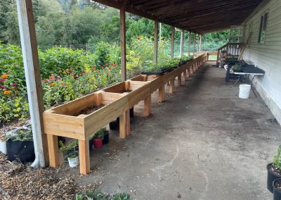 A line of raised wooden garden beds underneath a covered porch. Off to the side in the garden you can see many flowers in full bloom.